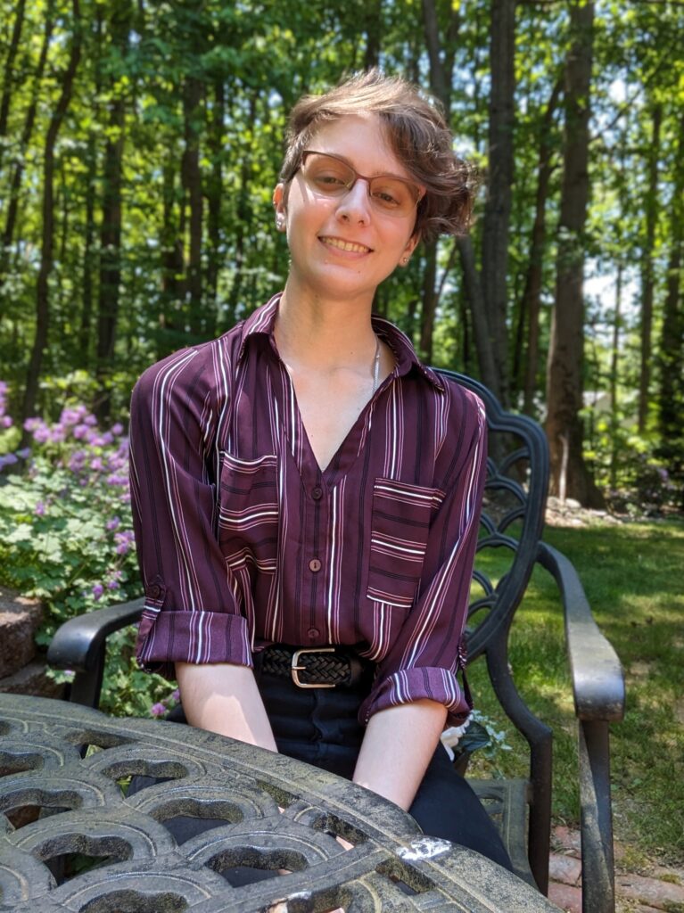 Portrait of a young white person with short hair and glasses sitting at an outdoor table